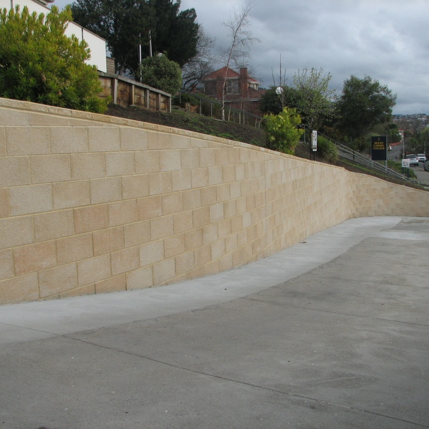 An expansive wall of Exposed Limestone follows the slope of a driveway on a suburban street, with overcast skies, trees, and houses creating a backdrop.