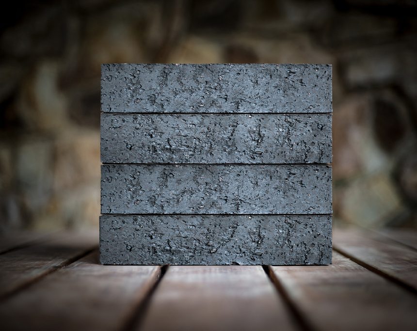 A stack of four "Slim Midnight" bricks, their deep Black hue evocative of the nighttime sky, rests on a wooden surface against a blurred stone wall background. The bricks feature a rough texture and are evenly aligned.
