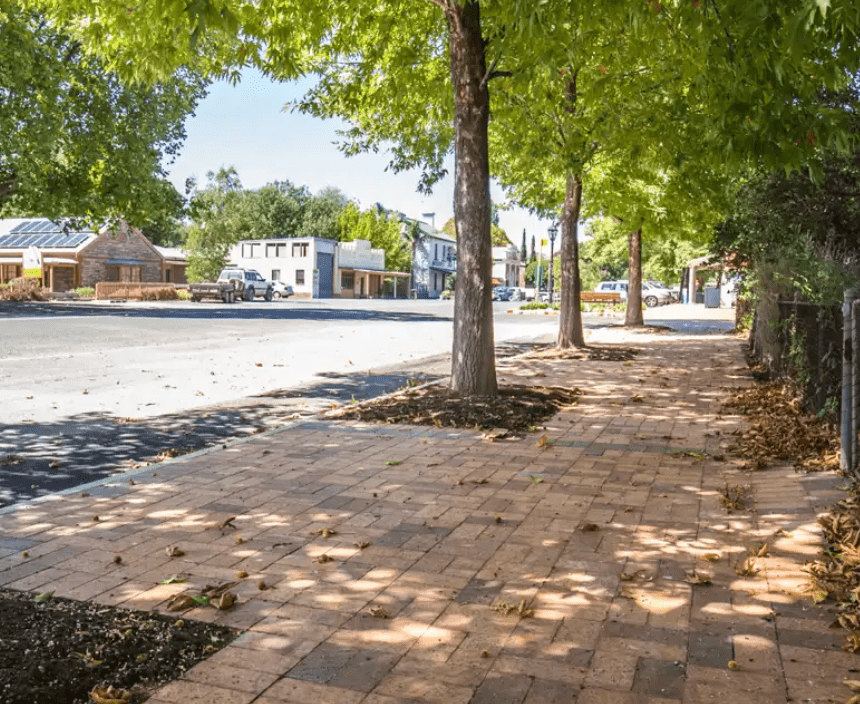 A tree-lined, brick-paved sidewalk basks in sunshine, with Hills Gold leaves on the ground. A quiet street runs parallel, flanked by charming houses and shops in the background, captured through a Hills Gold 50mm lens.