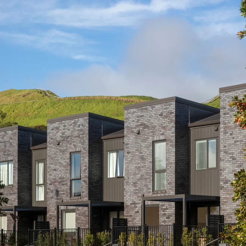 A modern row of dark-brick townhouses featuring large windows, small fenced yards, and scenic green hills—experience stylish living at La Palomo Azul.