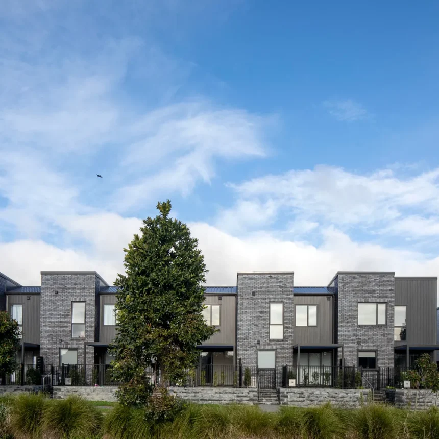La Palomo Azul is a modern row of gray brick townhouses with large windows and small front yards, set beneath a bright blue sky with scattered clouds, trees, and bushes in the foreground.