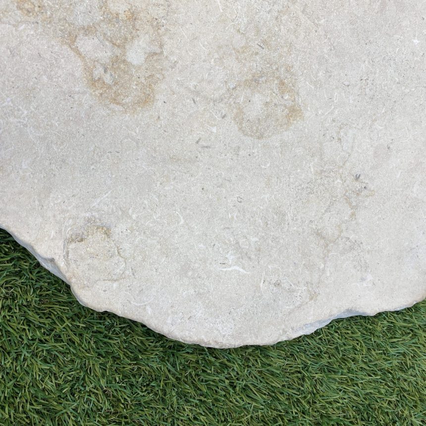 A large, flat stone, similar to the Crema Novelda Sandblasted & Tumbled Stepping Stones, features a rough, light grey surface as it lies on a bed of green grass. Its uneven edges create an elegant contrast against the neatly trimmed lawn beneath.