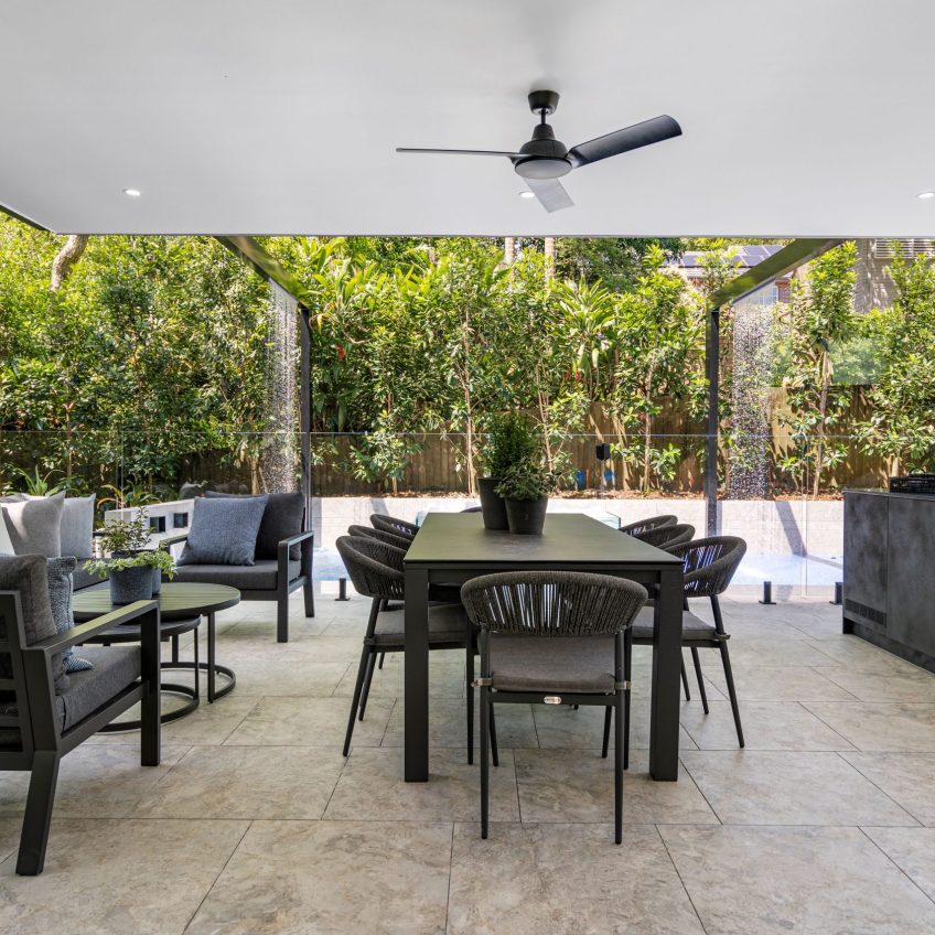 A contemporary outdoor patio showcases a dining table with chairs and cozy seating arrangements, all set on Multi Grey Grip Vit tiles. Above, a ceiling fan provides a gentle breeze, while the lush green garden in the backdrop enhances the serene and inviting atmosphere.
