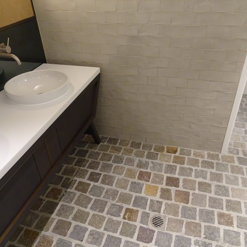 A bathroom corner features a white ceramic sink atop a dark wooden cabinet, set against a light beige tile-textured wall. The floor, adorned with Porphyry Filletti Split Edge random tiles in varying shades of grey and beige, elegantly complements the central metal drain.