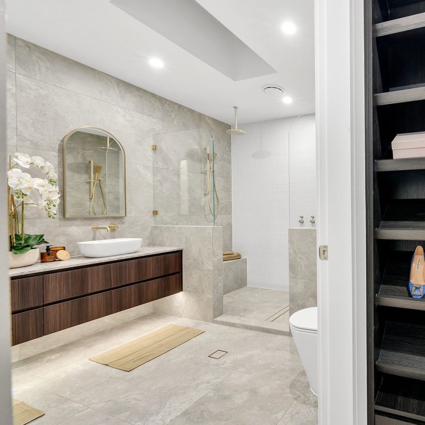 This contemporary bathroom highlights a stylish aesthetic, with a floating wood vanity paired with a white sink. A spacious mirror enhances the ambiance of the walk-in shower, which is adorned with Travertine Ocean Grip Vit tiles and features a rain showerhead. Orchids and decorative items contribute to the elegance of this peaceful retreat.