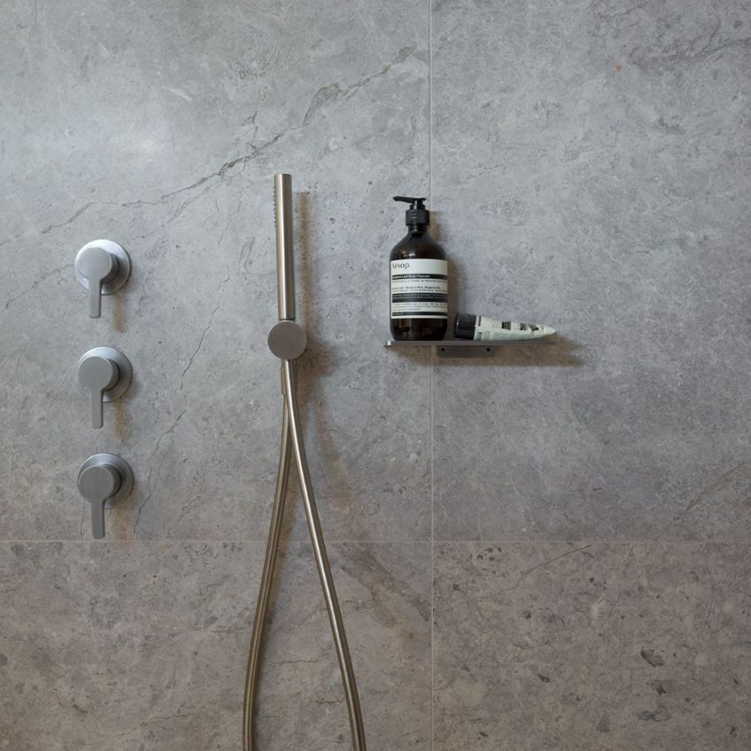 Featuring a modern shower with a sleek silver showerhead and three round control knobs, the Tundra Grey Sandblasted Tile 15mm wall provides an elegant backdrop. A small shelf neatly accommodates a pump bottle and a tube of toiletries, achieving the perfect balance of style and functionality.