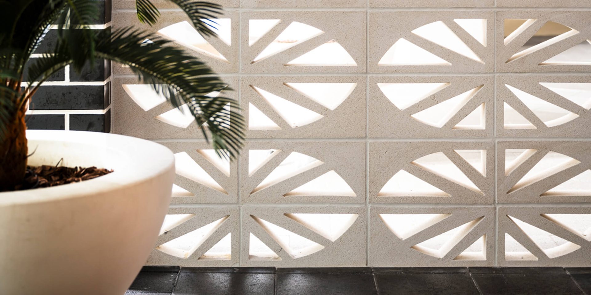 A wall made of Breeze Block Isabela features geometric cutouts that let light pass through. In front of it, a large white pot containing a leafy green plant is partially visible on the left side of the image. This design beautifully marries function and form.