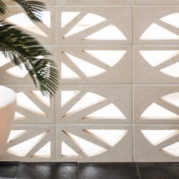A wall made of Breeze Block Isabela features geometric cutouts that let light pass through. In front of it, a large white pot containing a leafy green plant is partially visible on the left side of the image. This design beautifully marries function and form.