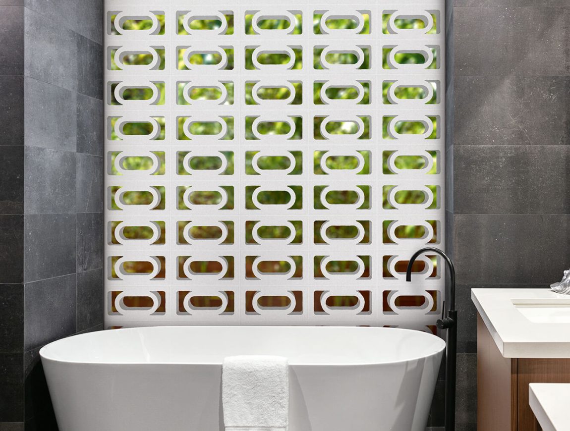 A contemporary bathroom featuring a white freestanding bathtub is complemented by dark grey tiled walls. The striking back wall showcases a pattern of green and white geometric tiles inspired by the Breeze Block Carnaval design. On the left, a black shower fixture adds contrast, while a white towel elegantly drapes over the tub's edge.