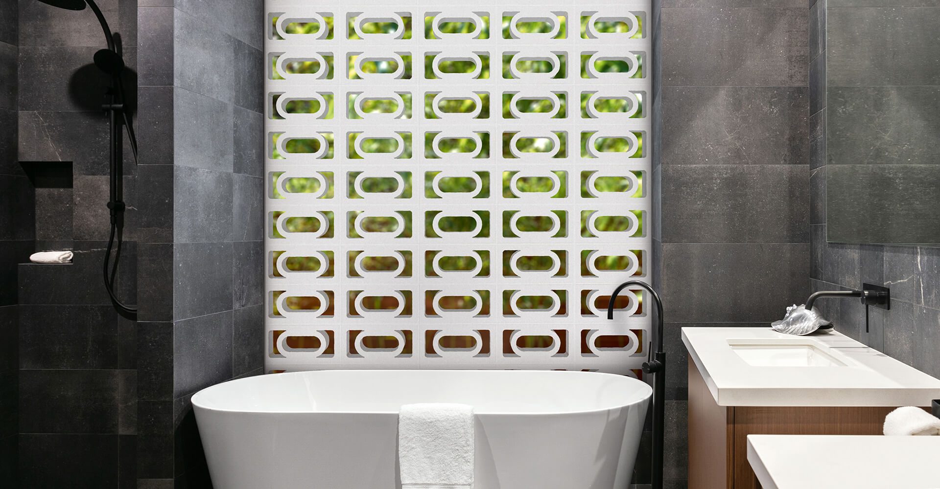 A contemporary bathroom featuring a white freestanding bathtub is complemented by dark grey tiled walls. The striking back wall showcases a pattern of green and white geometric tiles inspired by the Breeze Block Carnaval design. On the left, a black shower fixture adds contrast, while a white towel elegantly drapes over the tub's edge.