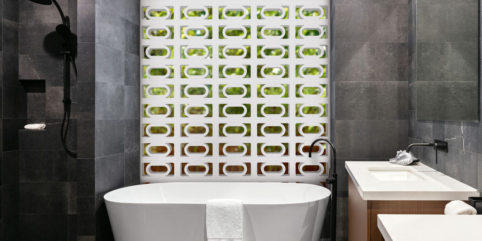A contemporary bathroom featuring a white freestanding bathtub is complemented by dark grey tiled walls. The striking back wall showcases a pattern of green and white geometric tiles inspired by the Breeze Block Carnaval design. On the left, a black shower fixture adds contrast, while a white towel elegantly drapes over the tub's edge.