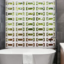 A contemporary bathroom featuring a white freestanding bathtub is complemented by dark grey tiled walls. The striking back wall showcases a pattern of green and white geometric tiles inspired by the Breeze Block Carnaval design. On the left, a black shower fixture adds contrast, while a white towel elegantly drapes over the tub's edge.