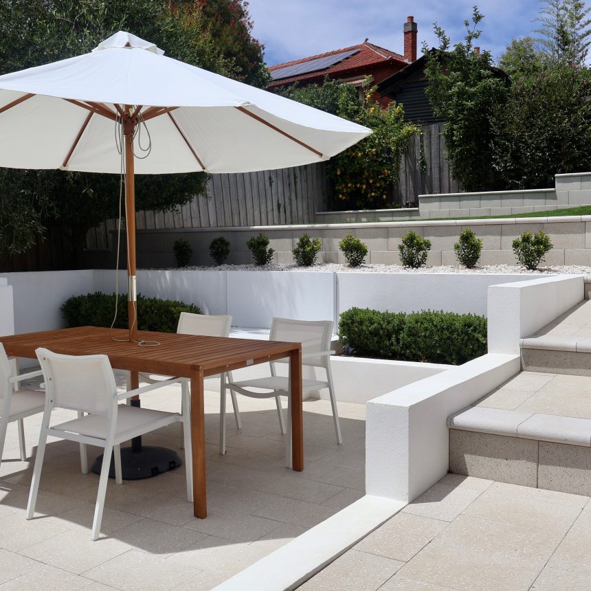 An outdoor entertaining area that is paved with light pavers and has and wooden table in the middle with four white chairs and a white umbrella through the table and a set of stairs on the right going up to a retained area