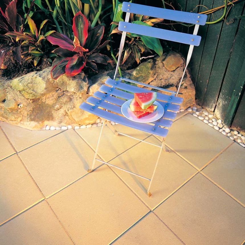 A blue wooden chair on the patio, near an Esplanade 400 Sand garden filled with green plants and rocks, holds a white plate with sliced watermelon. The light brown tiles perfectly complement the tranquil setting.