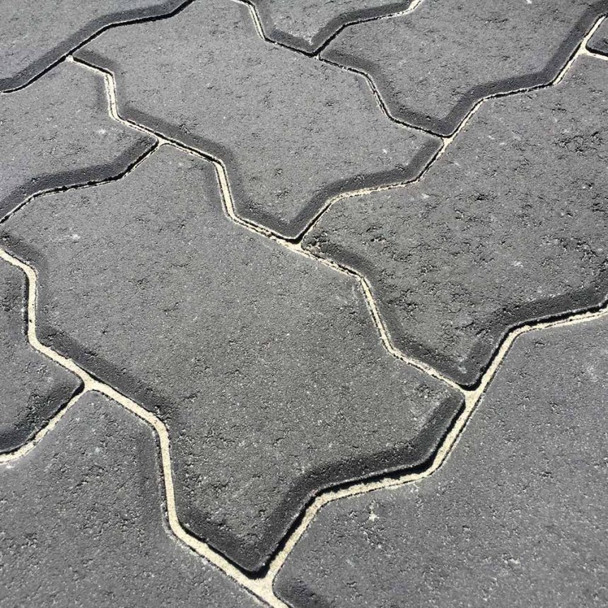 A close-up view of Interpave 80 Charcoal concrete pavers showcases their interlocking zigzag pattern. The surface looks slightly weathered, with light sand filling the gaps between the stones to provide stability.