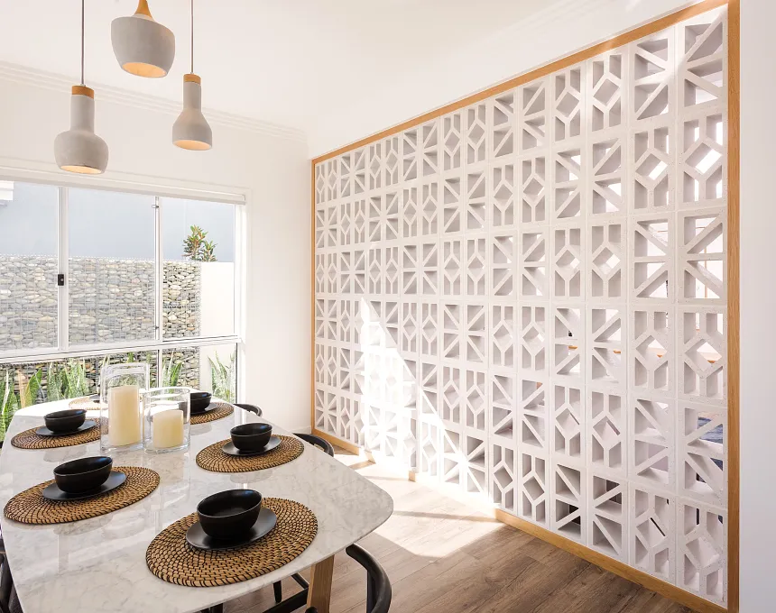 A dining area features a marble table for four, woven placemats, black dishware, three pendant lights, and a decorative GB Breeze Blocks Wedge Nickel screen wall that lets in natural light.