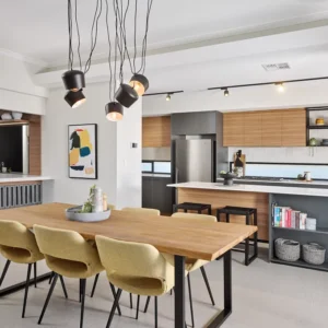 Modern kitchen and dining area with a wooden table, six beige chairs, pendant lights, kitchen island, and stainless steel appliances. Open shelving displays the GB Breeze Blocks Linear pewter completing the bright and spacious interior.