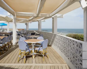 Outdoor dining area featuring blue and white striped chairs, wooden tables, and GB Breeze Blocks Diamond Porcelain accents on the deck, all shaded by a canopy with ocean views under a partly cloudy sky.