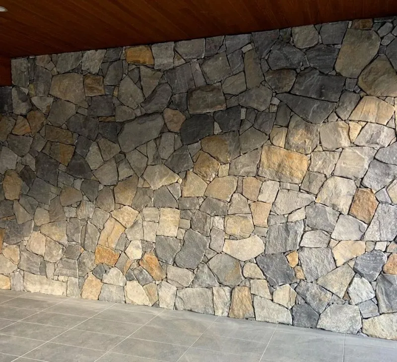 The image displays a wall crafted from irregularly shaped, variously sized grey and brown stones, echoing the artistic flair of the Freestyle Geneva design. The stones are skillfully arranged for a rustic look, complemented by a smooth, light grey tiled pattern on the floor.