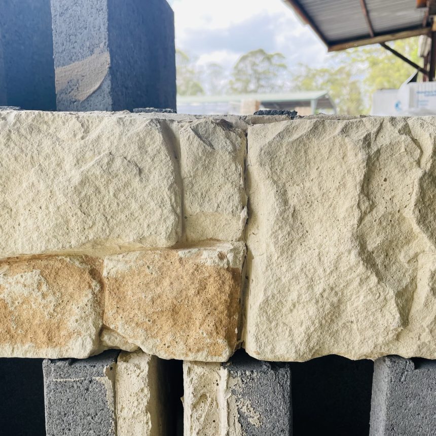 A stack of textured, sandy-colored StoneFace Bushrock bricks stands on the construction site. The background features other materials and a partially visible roof, with trees and a cloudy sky in the distance, framing this scene with rugged charm.