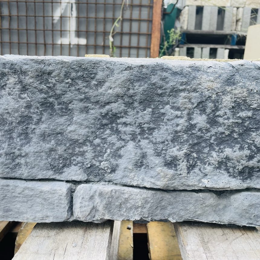 A sizable, rectangular block of StoneFace Bushrock in a gray hue is placed on a wooden pallet. Its textured surface and natural patterns showcase a distinct horizontal seam near the bottom, surrounded by industrial materials and additional pallets.
