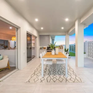 A modern outdoor dining area features a wooden table with six chairs and potted plants in GB Breeze Blocks Flower Porcelain on a patterned rug. The covered space is lit with recessed lights and opens indoors via sliding glass doors.