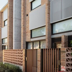 Contemporary brick townhouse with large windows, vertical white slats, concrete accents, and a modern metal gate. The GB Breeze Blocks Wedge Nickel adds geometric brickwork, while lush greenery completes the striking facade.