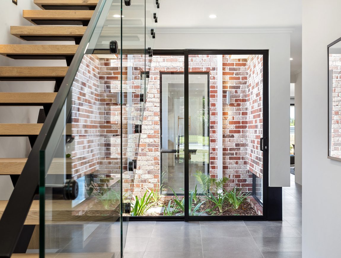 Modern interior featuring a wooden staircase with glass railing on the left. A windowed wall looks out onto a small courtyard with brick walls crafted from Midland Bricks and lush plants. The space is bright and minimalistic, with gray floor tiles and simple lighting.