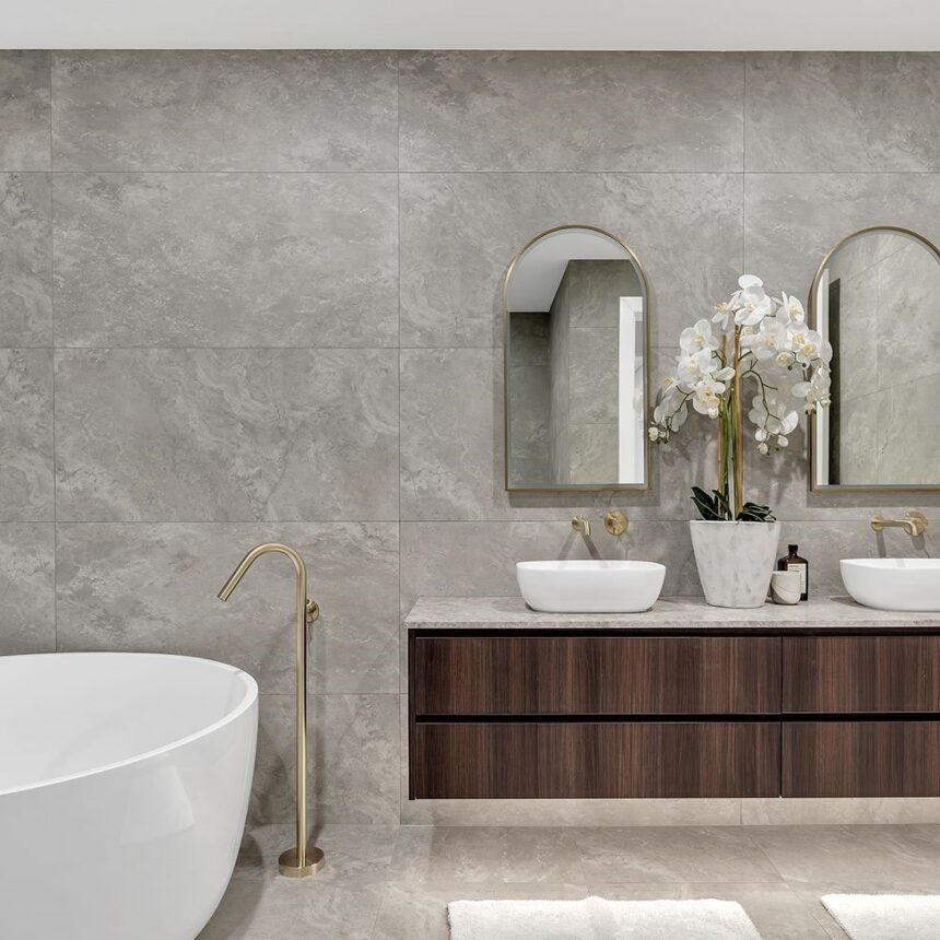 Luxurious bathroom with gray tiled walls and a Travertine Ocean Grip Vit floor. Includes a white freestanding tub, dual sinks on a wooden vanity, two oval mirrors, and a vase of white orchids. Natural light pours in through a small window.