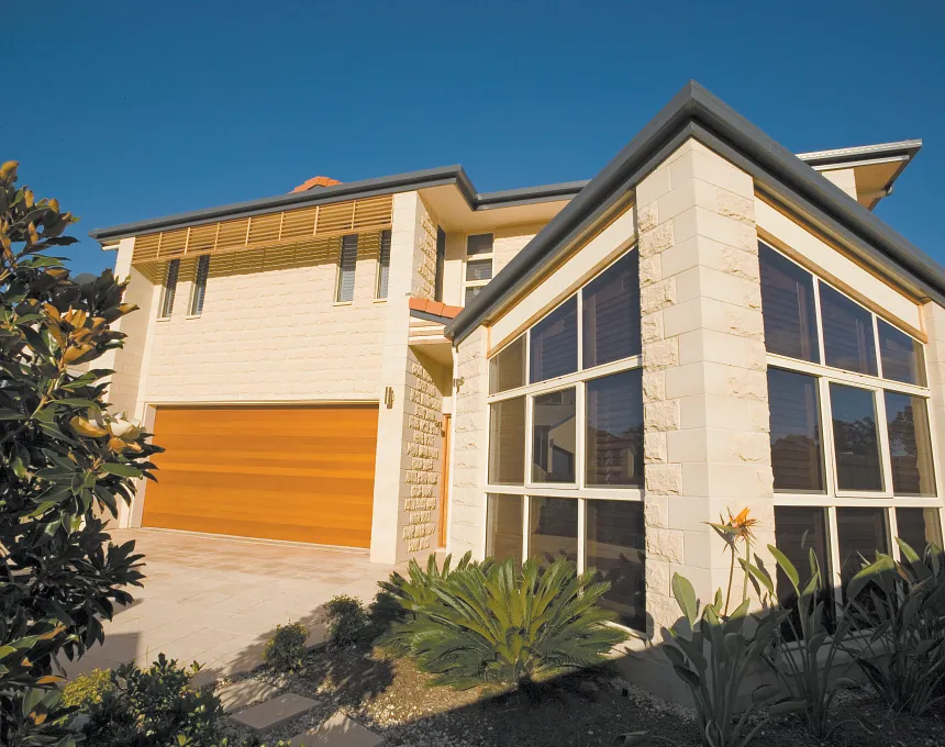 Modern two-story house with large windows, GB Sandstone Rockface Limestone exterior, wooden garage door, sloped roof, and landscaped garden beneath a clear blue sky.