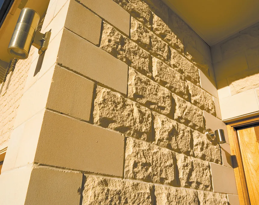 Close-up of a building corner featuring GB Sandstone Rockface Limestone blocks with a textured finish, smooth rectangular panels, two metallic wall lights, and a glimpse of a wooden door on the right.