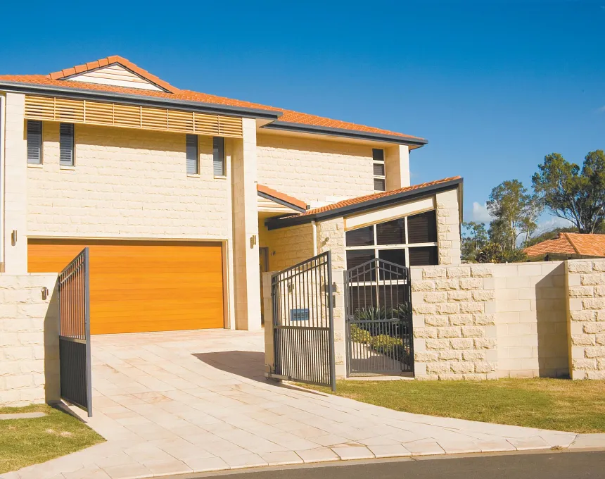 Modern two-story house features GB Sandstone Rockface Limestone walls, a spacious wooden garage door, black metal gate, and paved driveway. Red-tiled roof enhances the landscape of green lawn and trees.