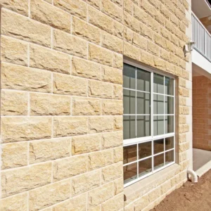 A close-up of a building exterior showcases large windows, a small balcony, and bare dirt beside the wall, all finished with GB Sandstone Rockface Sydney Blend bricks in light tan.