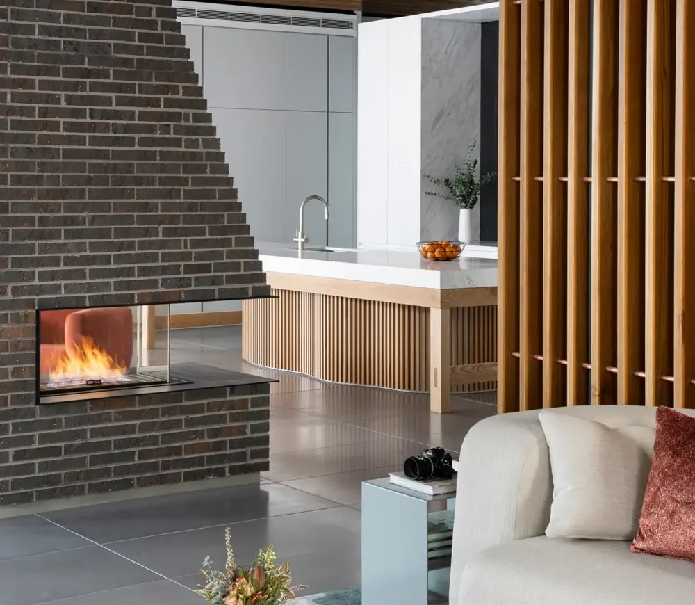 The Long Coachhouses modern living room features a brick fireplace on the left, a white sofa with a rust pillow on the right, and in the background, a sleek kitchen showcasing a wooden island with a silver faucet. A small vase of flowers graces the table in front.