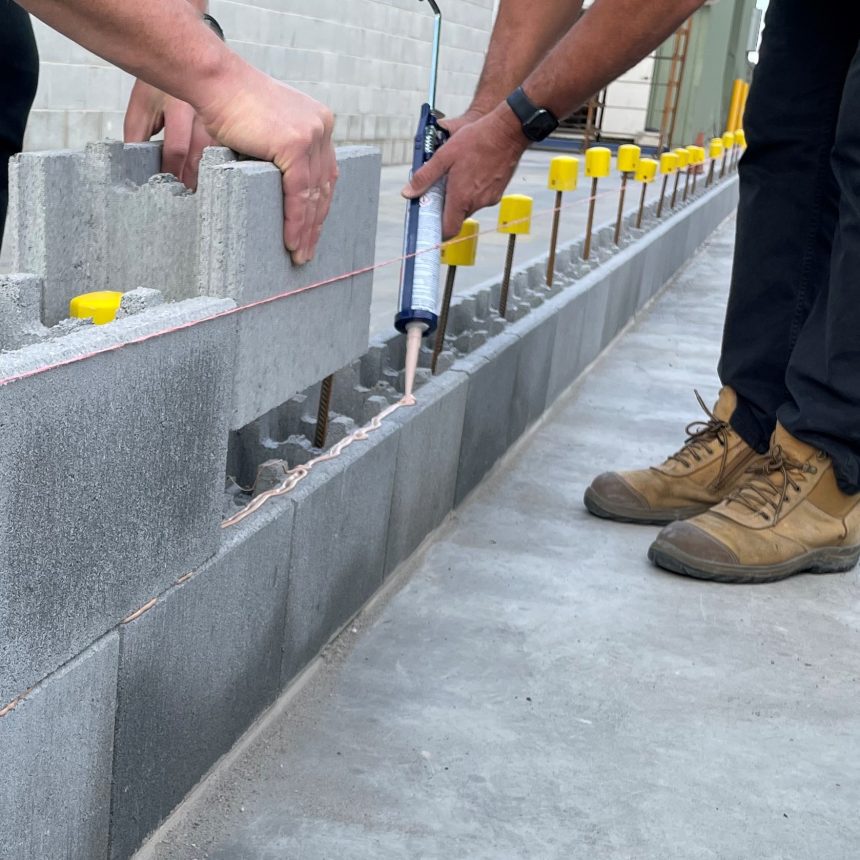Two workers in construction clothes lay DIY Coloured Blocks Mist ECO in a straight line. One applies adhesive with a caulking gun, while the other positions the blocks. A string line guides alignment, complemented by yellow spacers in the background, showcasing precision and DIY flair.