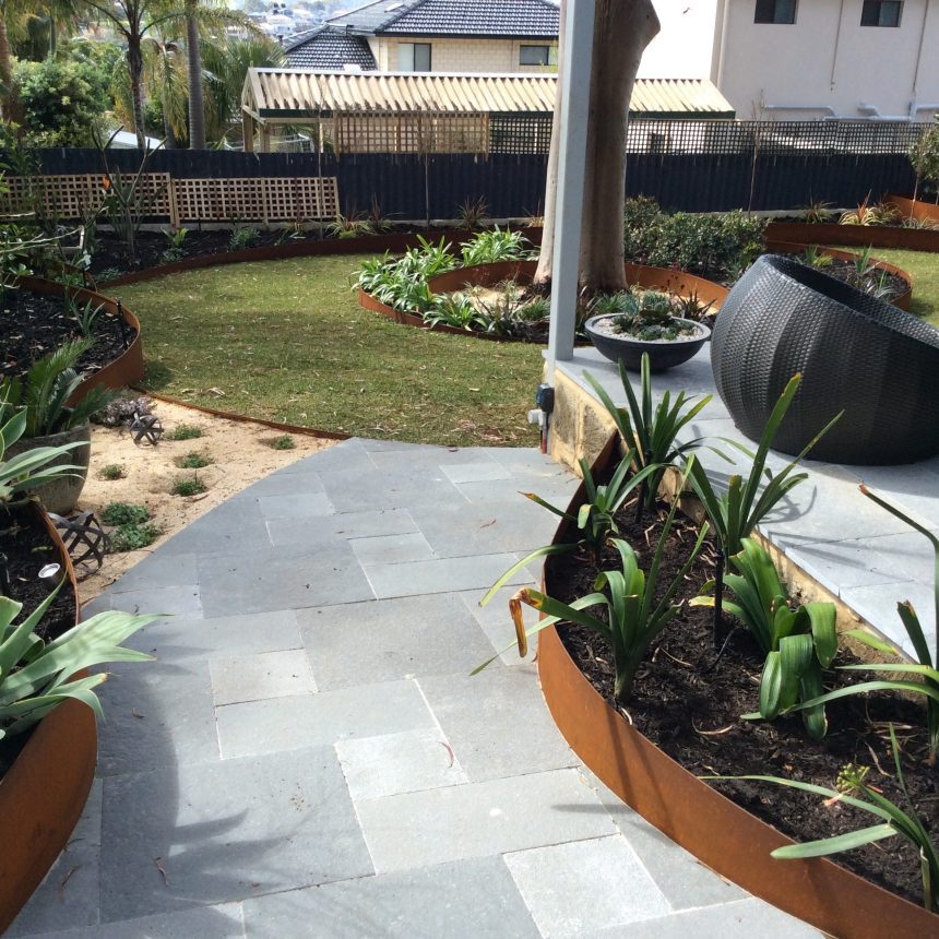 A beautifully landscaped modern backyard garden features a stone pathway leading to a patio. Circular planters with lush green plants complement the outdoor seating. The well-maintained lawn is framed by fences, with neighboring houses visible in the background.