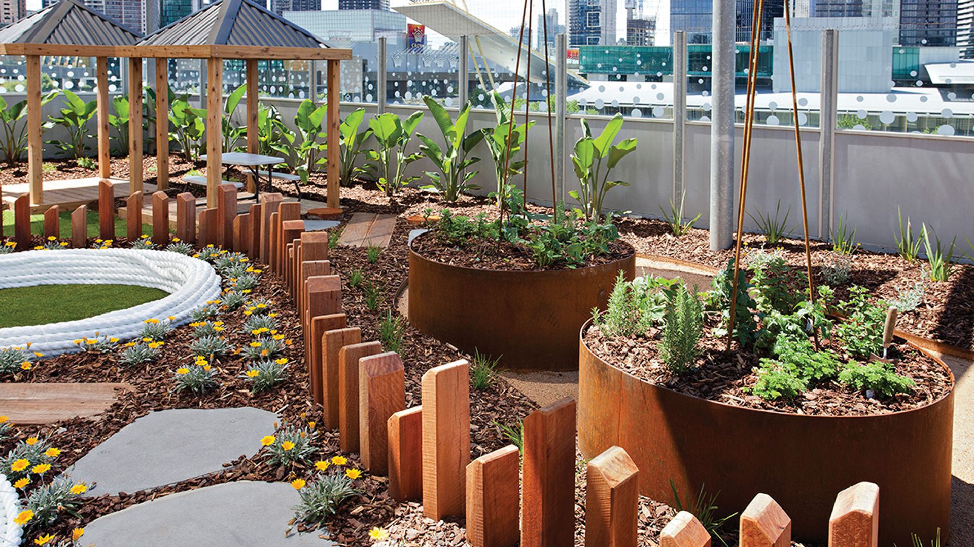 A rooftop garden features large round planters filled with greenery, bordered by FormBoss edging and vibrant yellow flowers. A small gazebo and stepping stones enhance the space, all set against a striking city skyline backdrop.