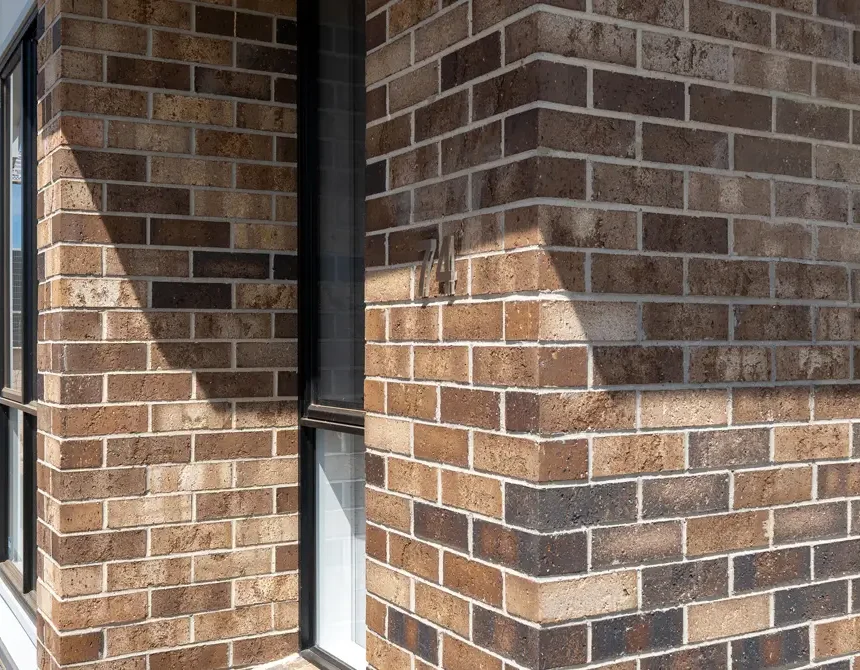 The essence of the Murray Darling architectural style is captured in this close-up of a brick buildings corner, featuring brown and beige bricks. Sunlight casts shadows that highlight the texture, while a window on the left partially reflects light.