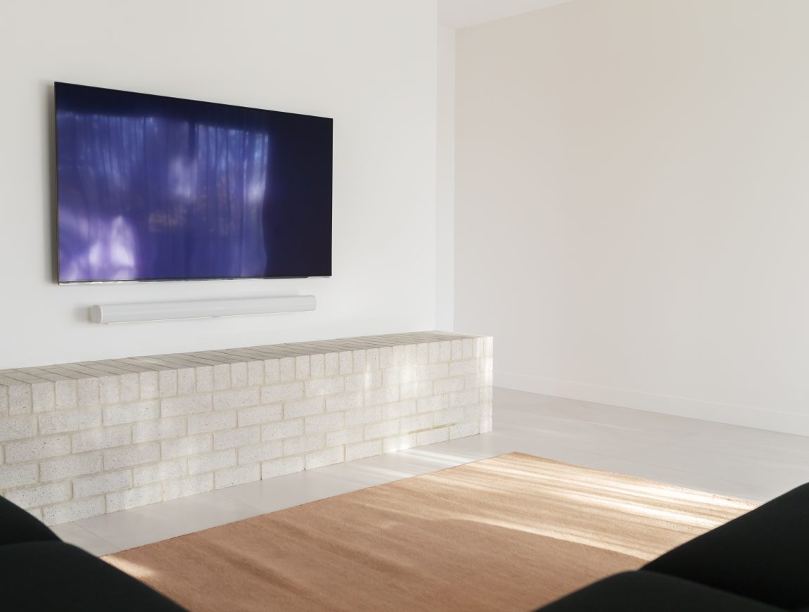 A modern living room showcasing internal design inspiration with a wall-mounted TV above a light brick ledge and soundbar. Minimalistic decor includes a soft brown rug on white tiled flooring, complemented by two black couches facing the TV.