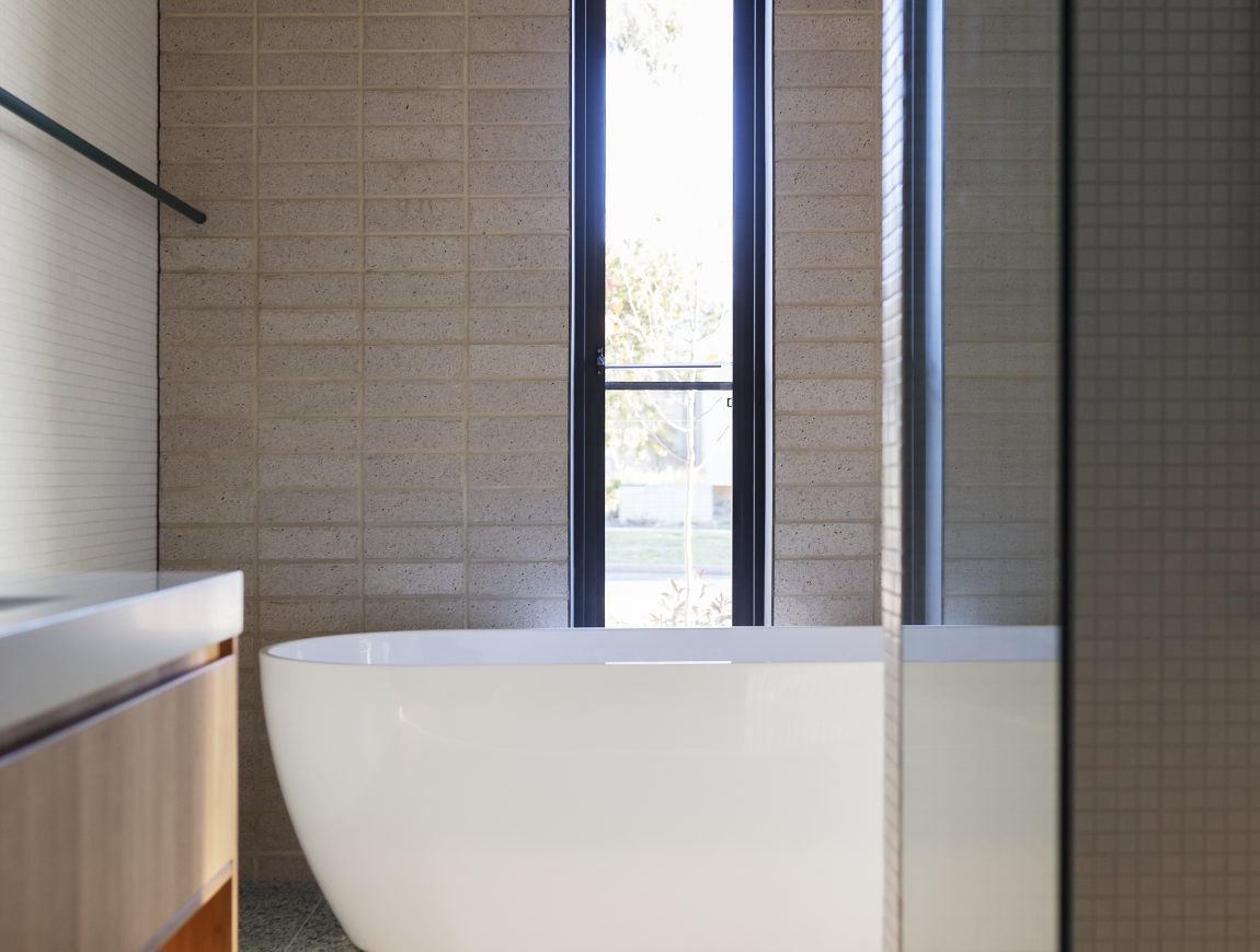 A modern bathroom offers internal design inspiration with its freestanding white bathtub. The space features a tall narrow window, a wooden ceiling, and a vanity with wooden accents. Speckled gray tiles cover the floor, enhancing the minimalist design.