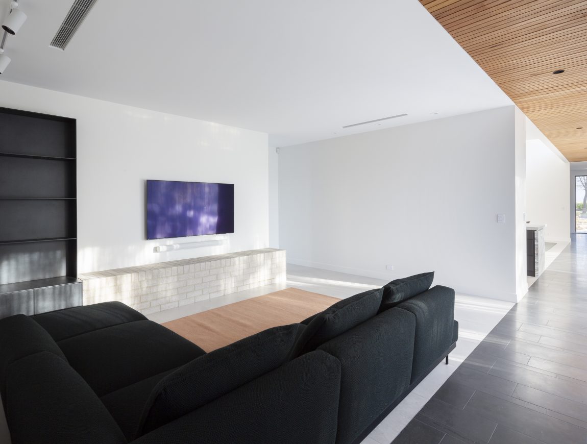 Modern living room with a minimalist design featuring a large black sofa, flat-screen TV mounted on a light brick wall, and an empty black bookshelf. This inspiration in internal design showcases a sleek wood ceiling panel and natural light streaming in from a hallway.