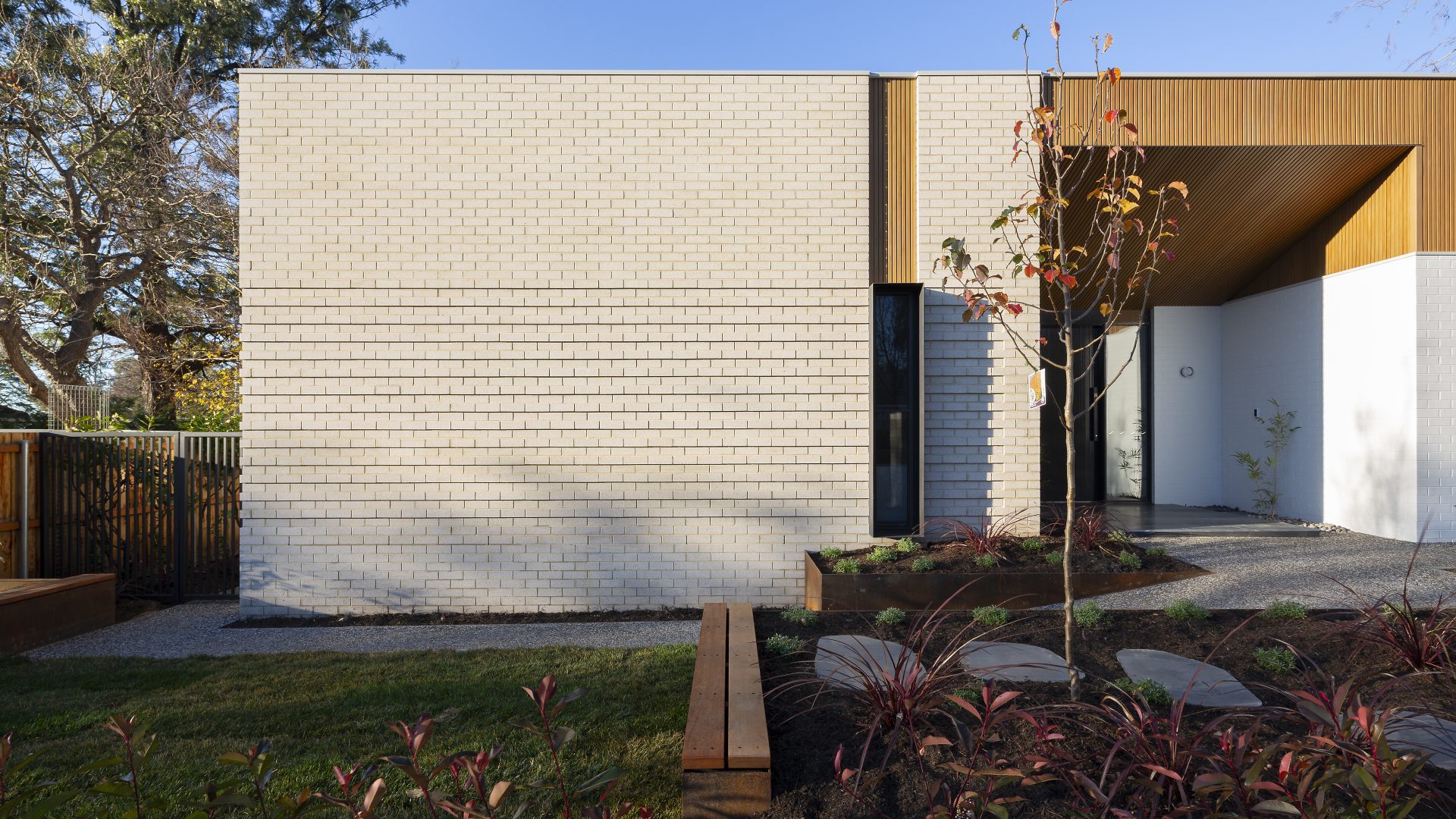 Modern single-story house with light brick exterior. A pathway leads to the entrance, surrounded by a landscaped garden with sparse plants and a young tree. Drawing internal design inspiration from nature, the setting features a wooden fence and tall trees against a clear blue sky.