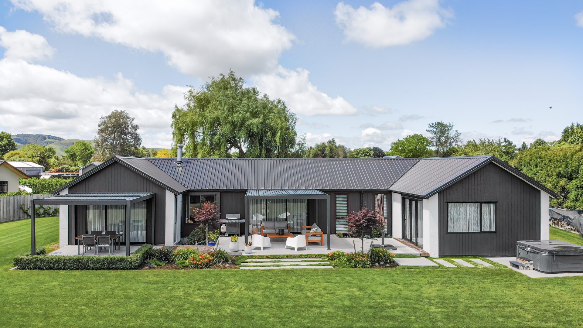 A modern, single-story black house with a metal roof offers design inspiration through its large windows and lush green lawn. An outdoor patio area features a gazebo, seating, and a barbecue. The serene backdrop includes trees and a partly cloudy sky.