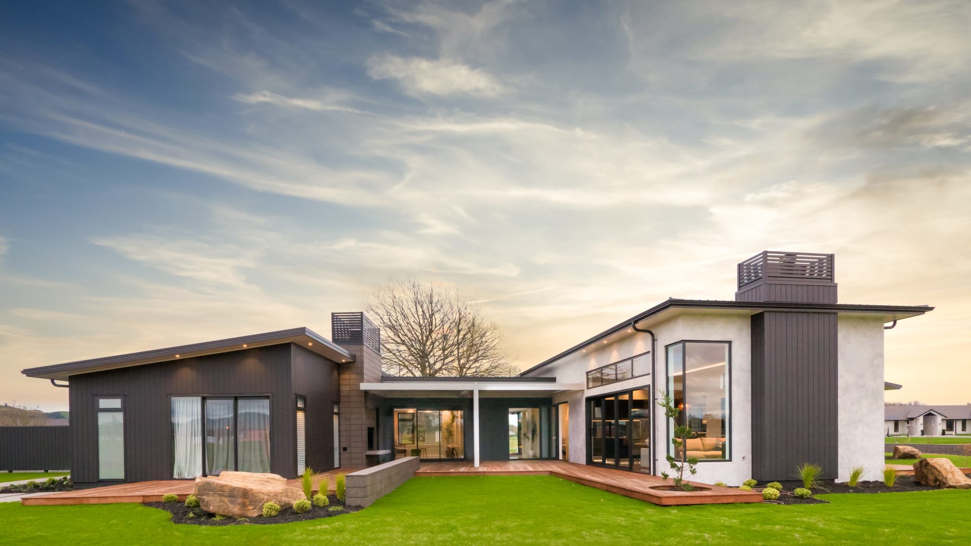 Modern single-story house with large windows and a flat roof, surrounded by a green lawn. The exterior features a mix of dark and light finishes. The sky is partly cloudy, adding a picturesque backdrop.