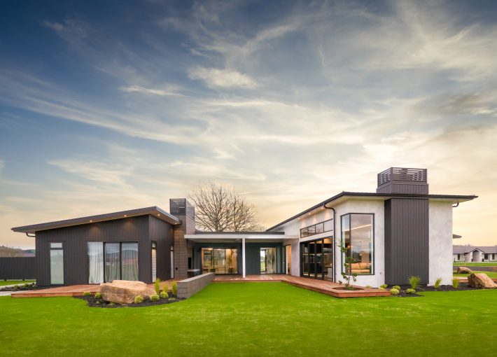 Modern single-story house with large windows and a flat roof, surrounded by a green lawn. The exterior features a mix of dark and light finishes. The sky is partly cloudy, adding a picturesque backdrop.