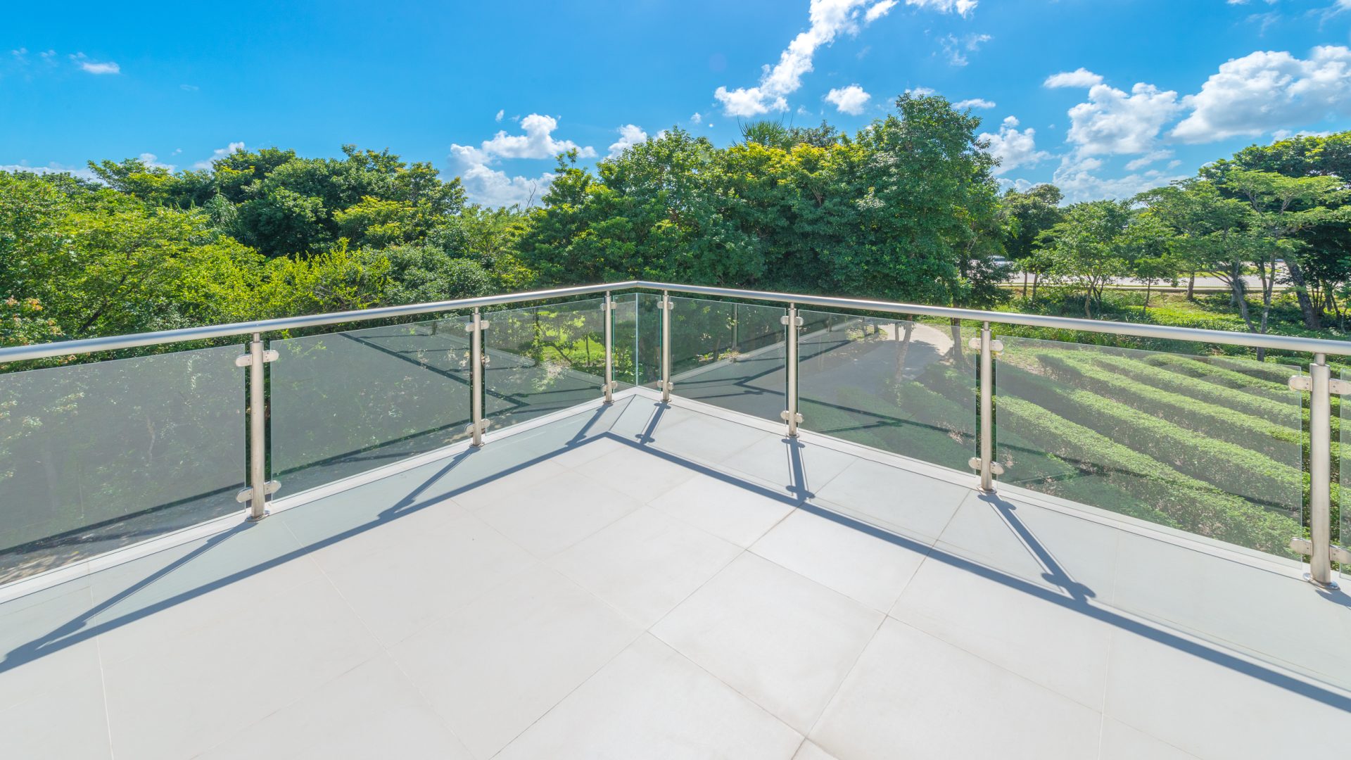 A sunny balcony with a Midland Brick railing overlooks a lush green landscape and trees. The clear blue sky and scattered clouds enhance the bright and serene atmosphere.