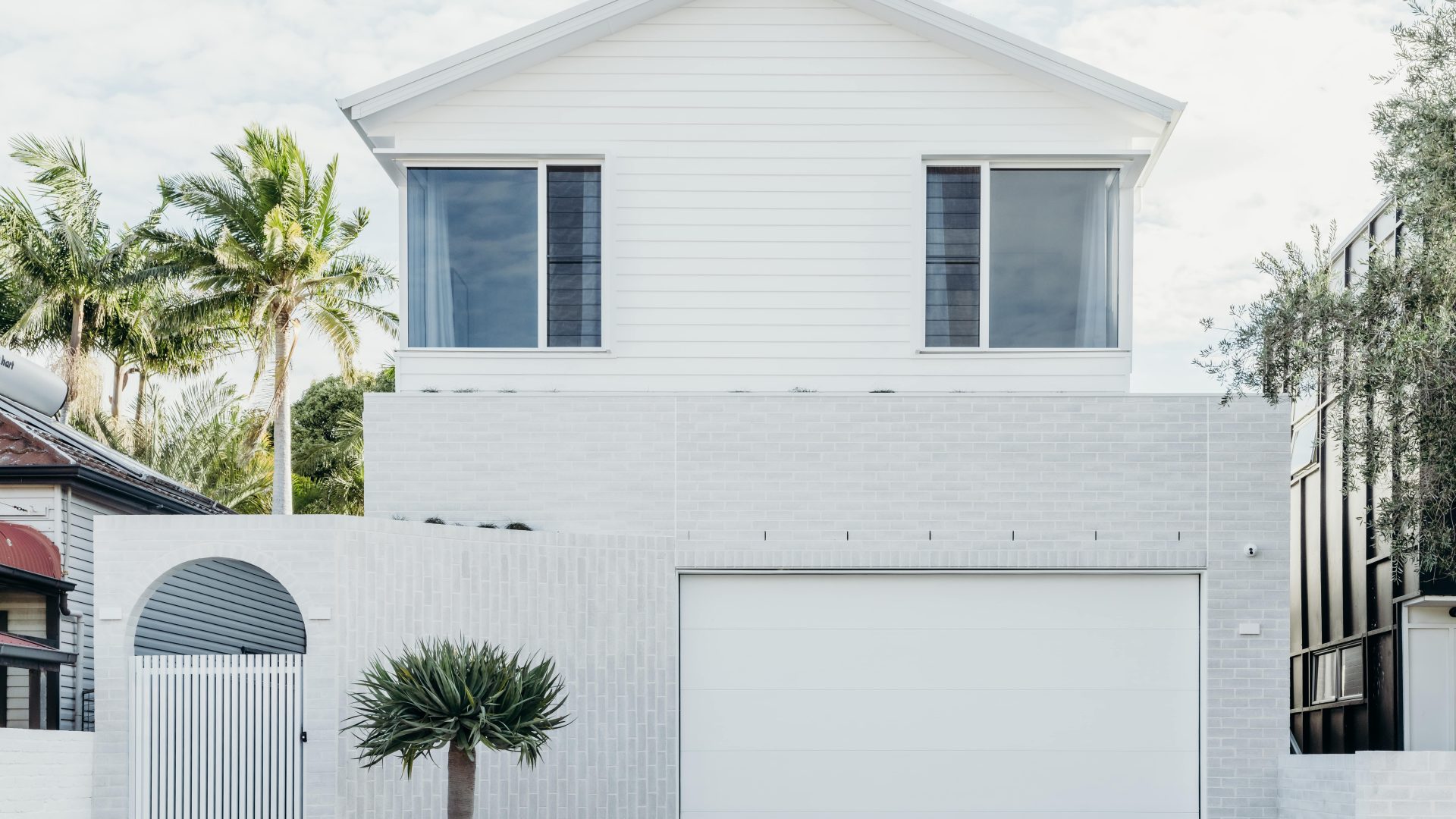 The modern white two-story house, featuring Architectural Designer Products, boasts a sleek exterior with minimalistic design elements and a large garage door. A small tree in a planter graces the entrance, while palm trees sway gently in the background.