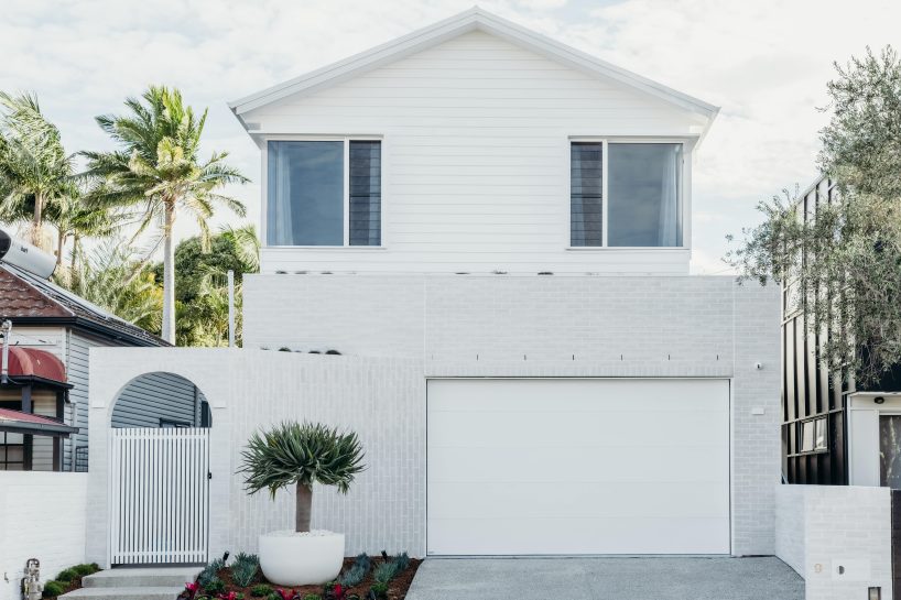 A modern two-story white house, showcasing Architectural Designer Products, features large windows, a white garage door, and a gated entrance. The front yard is adorned with a potted plant and palm trees beneath the partly cloudy sky.