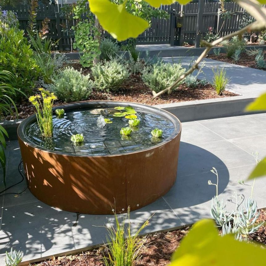 A garden with a round, rust-colored water feature containing water lilies is bordered by a gray paved path of Austral Bricks. Surrounding it are various plants and shrubs, set against a wooden fence, with sunlight filtering through tree branches.