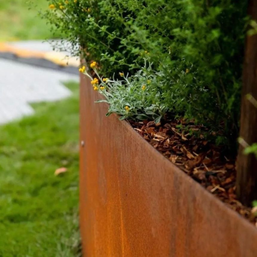 Close-up of a rust-colored metal planter from FormBoss filled with lush greenery and small yellow flowers. The planter sits in a grassy area, with a pathway visible in the background, slightly out of focus.